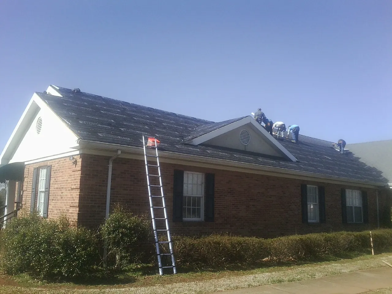 Crew tearing shingles off a brick ranch roof with a ladder set — Emergency Roof Repair work by our crew in Fawn Grove