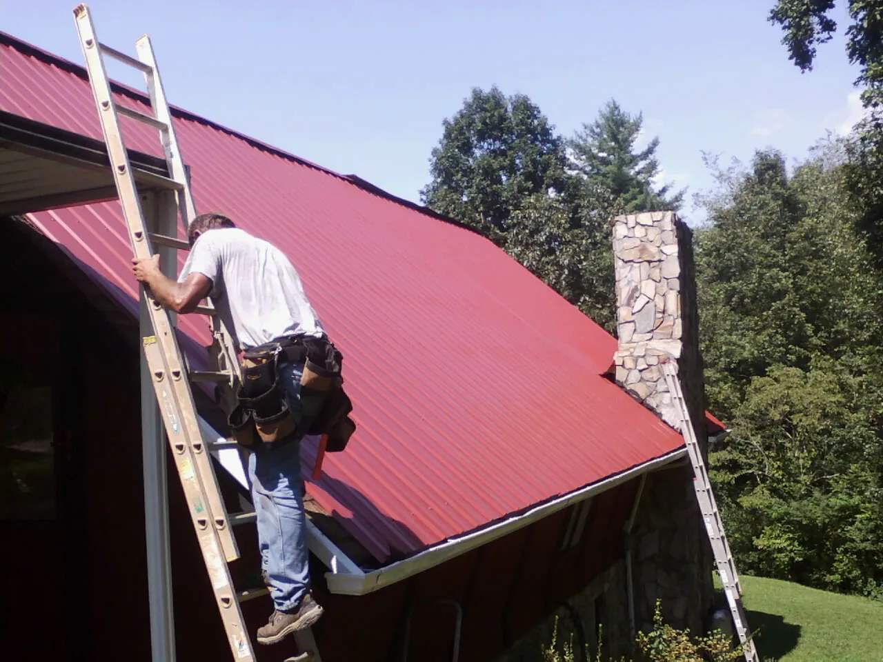 Roofer on a ladder beside a red metal roof and stone chimney — Soffit & Fascia Repair by Roofing Brothers in Fawn Grove